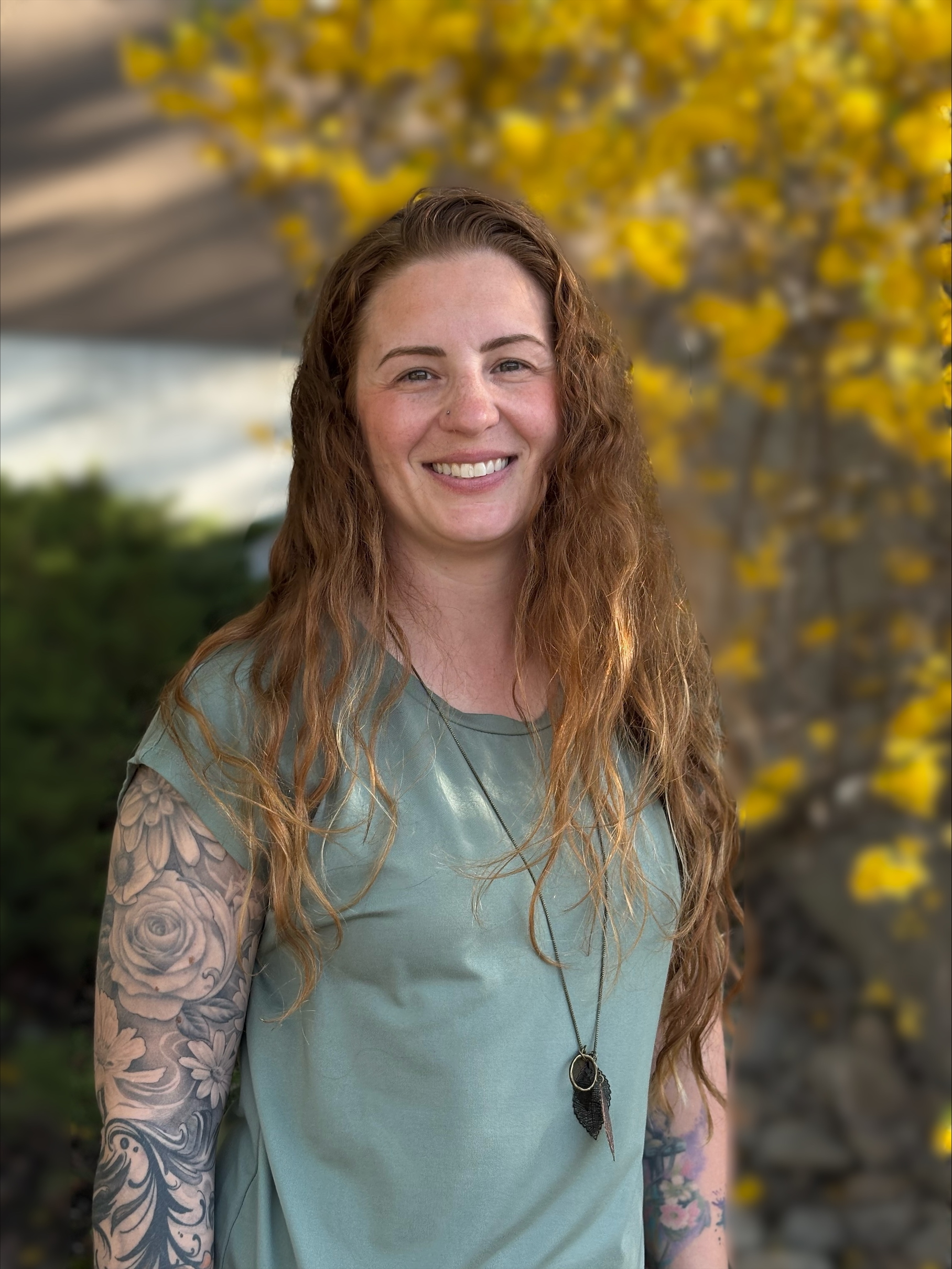 Smiling woman with long curly hair wearing a green t-shirt, a leaf pendant necklace, and a sleeve tattoo.