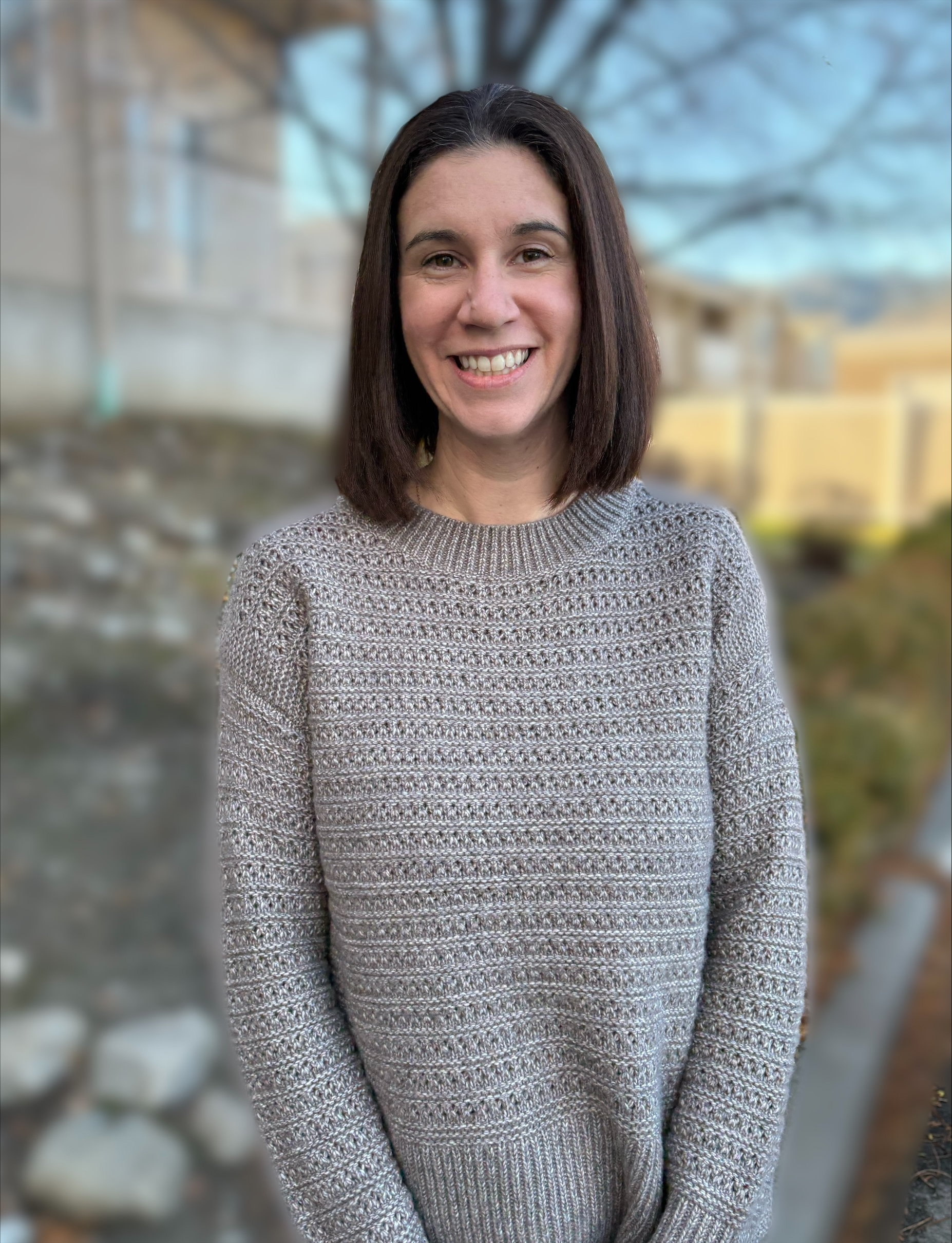 A smiling woman with shoulder-length brown hair, wearing a grey knit sweater in a bright, outdoor setting.