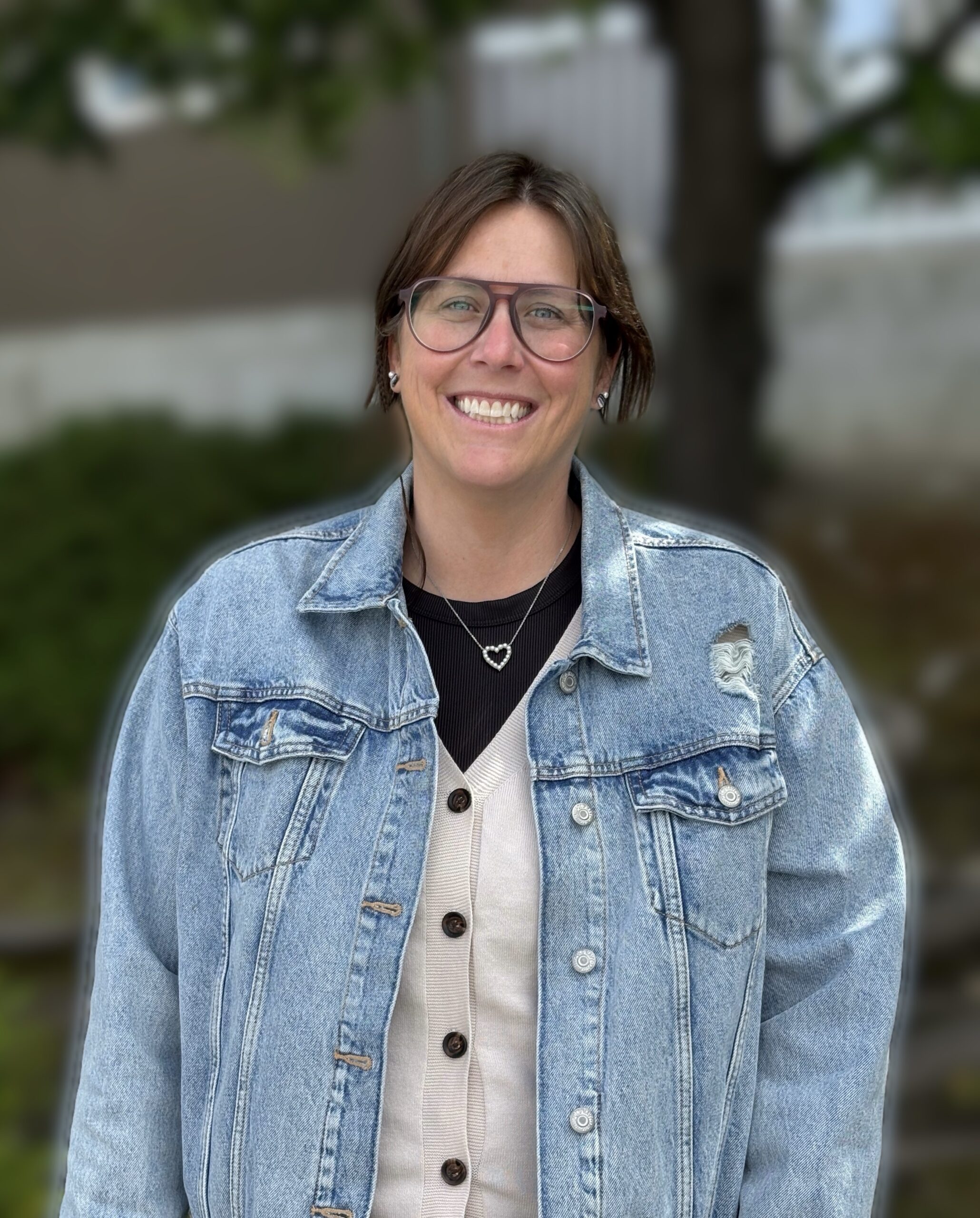 "A smiling woman with short brown hair and glasses, wearing a denim jacket over a beige cardigan and a black shirt with a heart necklace. She is standing outdoors with a blurred background of green trees.